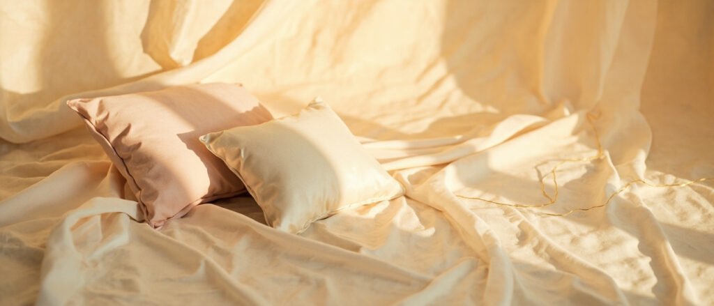 Coussin satin champagne drapé, plis ivoire, lumière dorée, espace à gauche.
